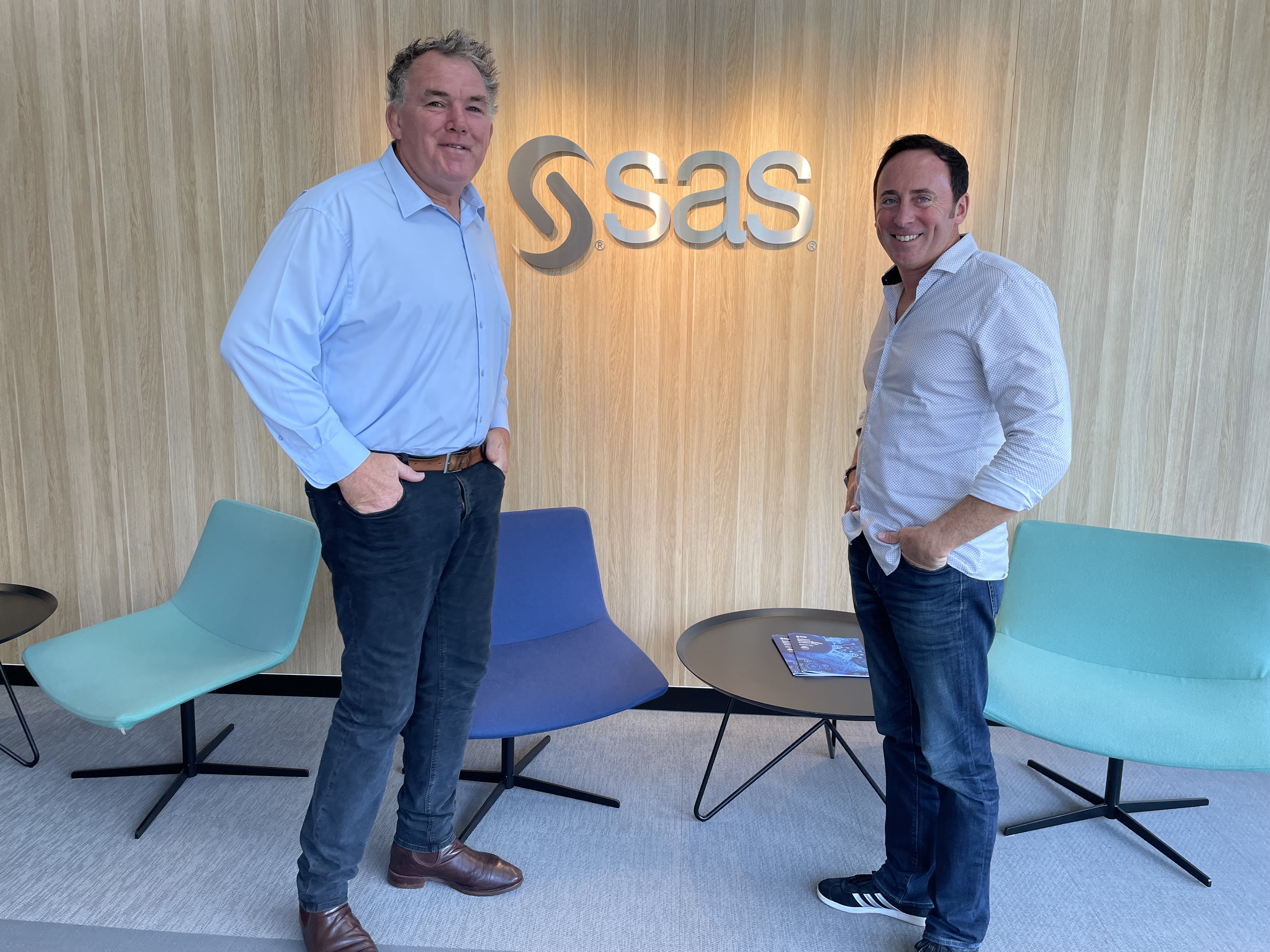 Owen Finegan and SAS Vice President Craig Jennings stand in front of some chairs and tables which are set up in front of a wood-panel wall featuring the SAS logo.