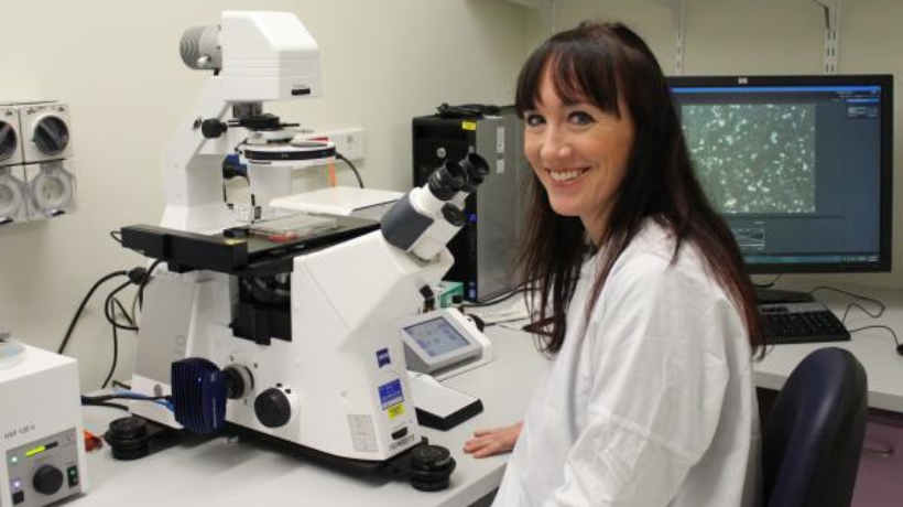 Dr Rachel Conyers in a lab gown, sitting in front of a microscope and other research equipment